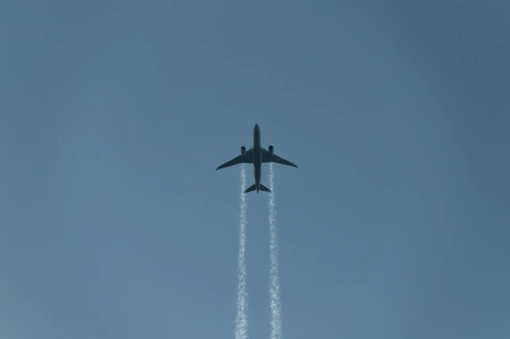 A commercial airplane leaving contrails as it flies across a clear blue sky, viewed from below.