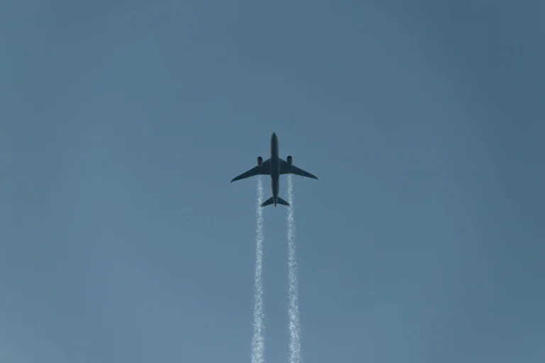 A commercial airplane leaving contrails as it flies across a clear blue sky, viewed from below.