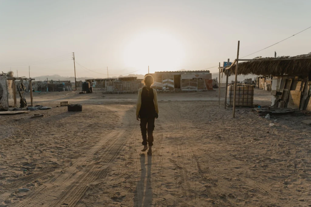 A solitary person walks through a desert settlement at sunrise, casting a long shadow on sandy terrain.