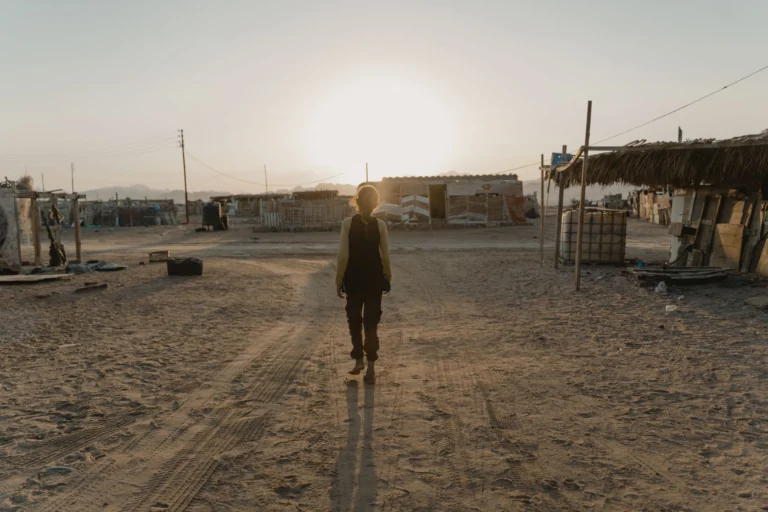A solitary person walks through a desert settlement at sunrise, casting a long shadow on sandy terrain.
