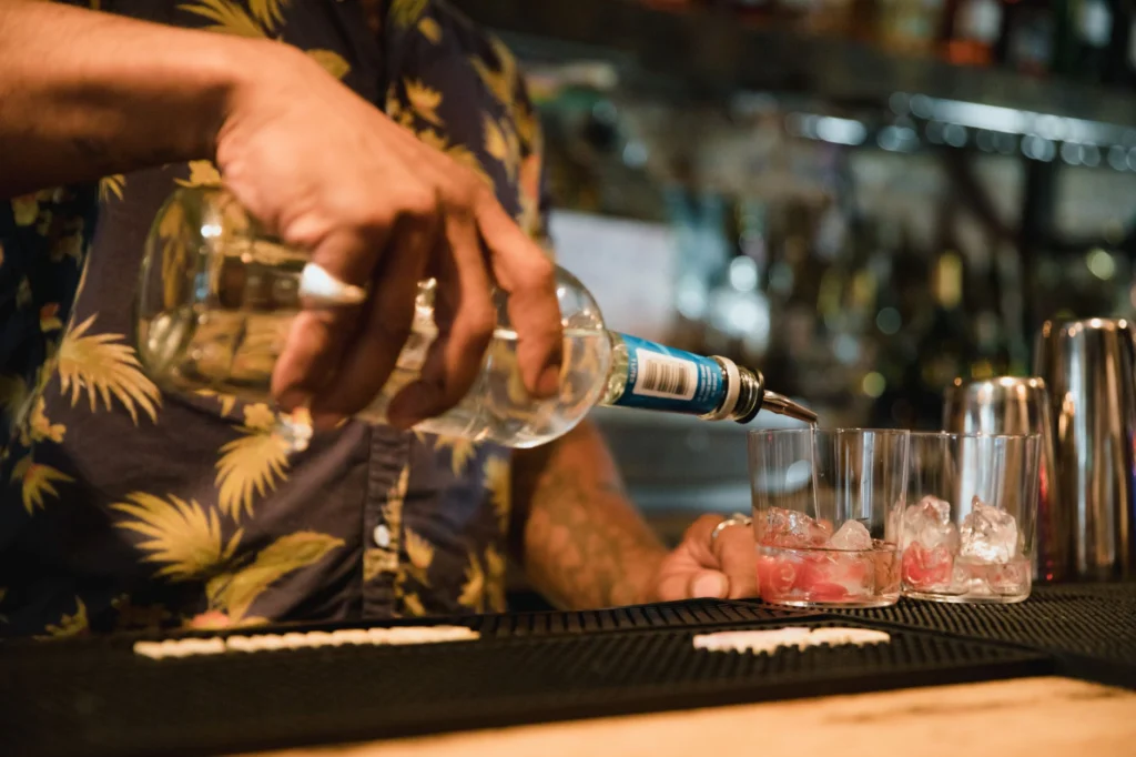 A bartender skillfully pours drinks into glasses filled with ice at a lively cocktail bar.