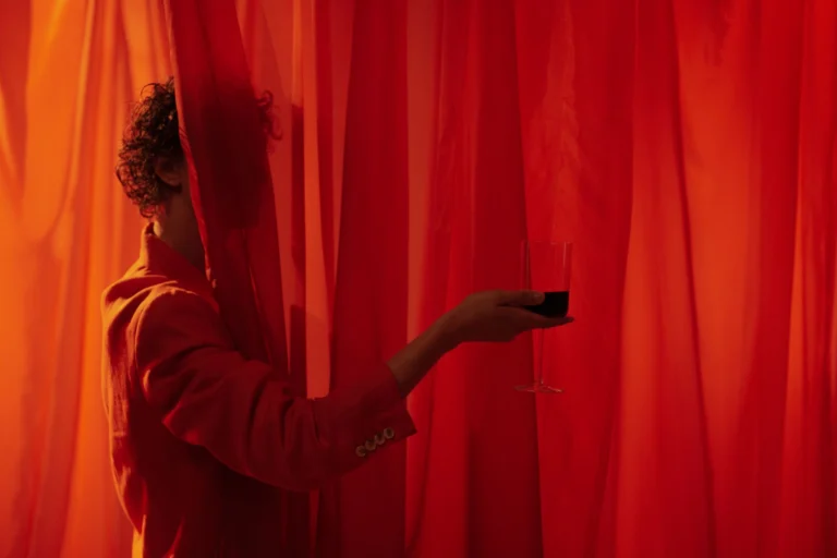 A mysterious figure in a red suit holds a glass of wine behind vibrant red curtains.