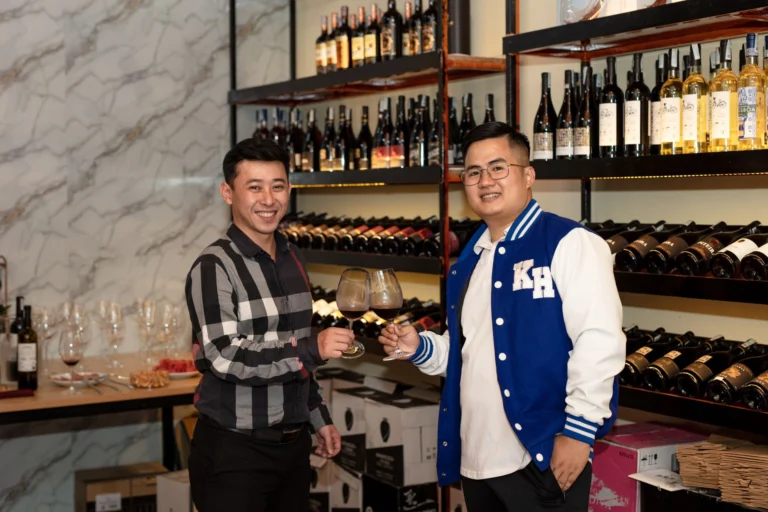 Two Asian men toast with wine glasses in a cozy wine cellar setting, surrounded by bottles.