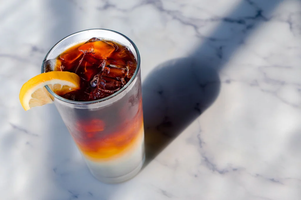 Tall glass of iced tea with lemon slice on a sunny marble surface casting a shadow.