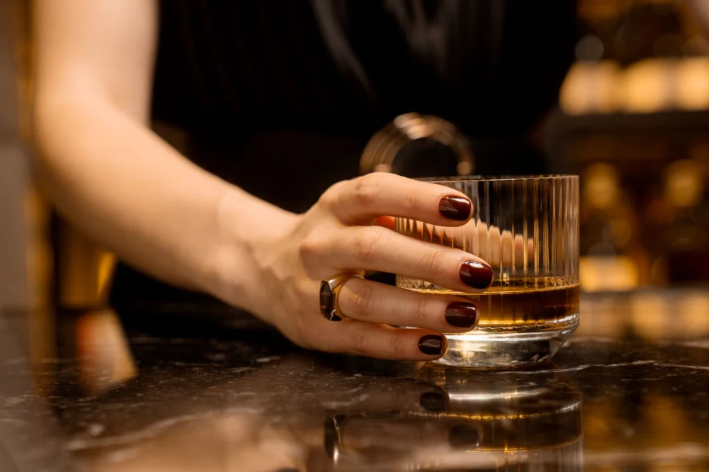 Close-up of a woman's hand with manicured nails holding a whiskey glass on a marble bar top.