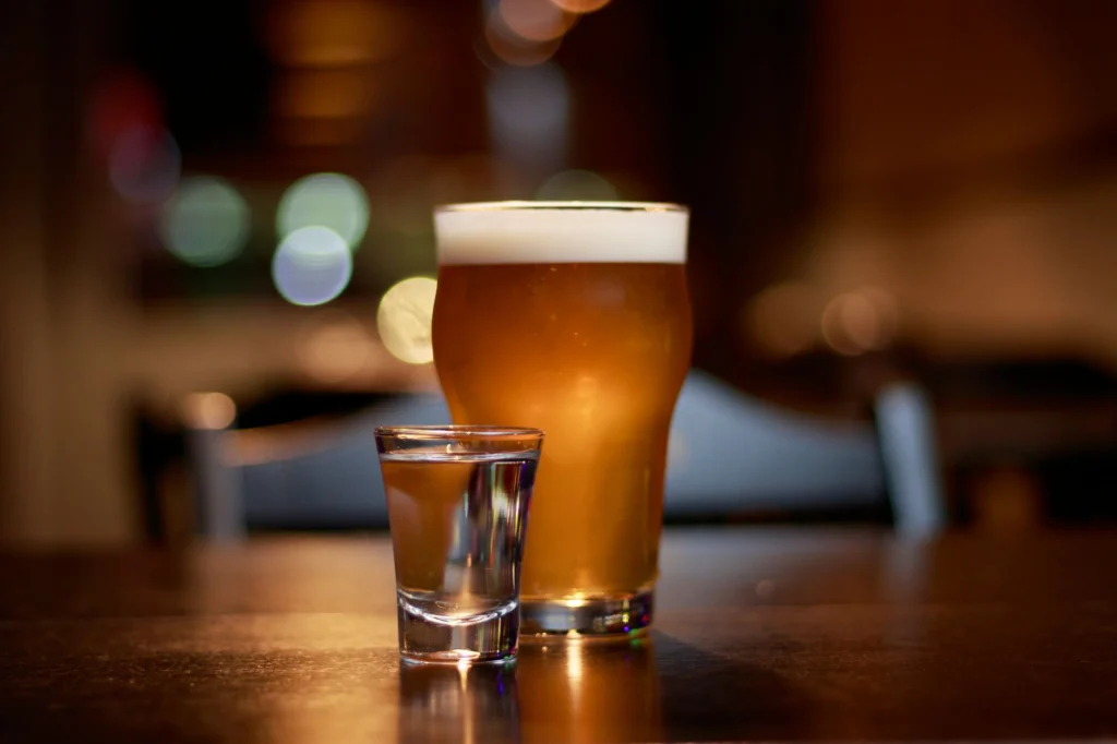A close-up of a beer glass and vodka shot on a pub table with bokeh lighting.