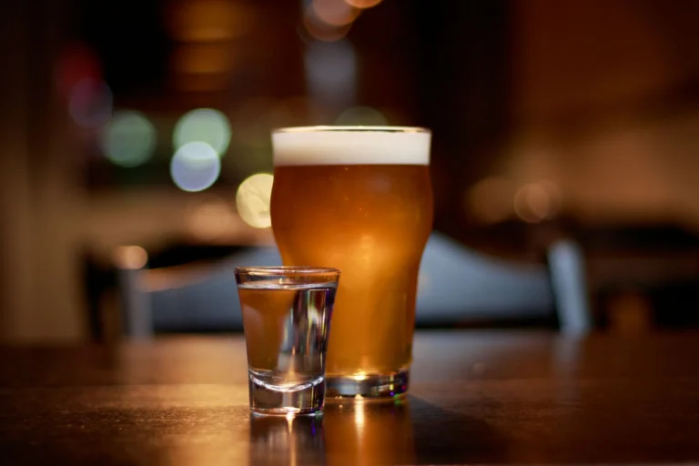 A close-up of a beer glass and vodka shot on a pub table with bokeh lighting.