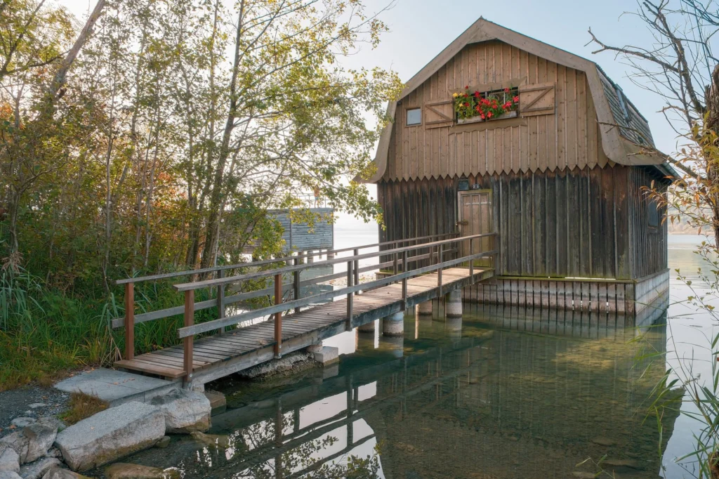 Wooden house by lake with a rustic bridge and colorful flowers.