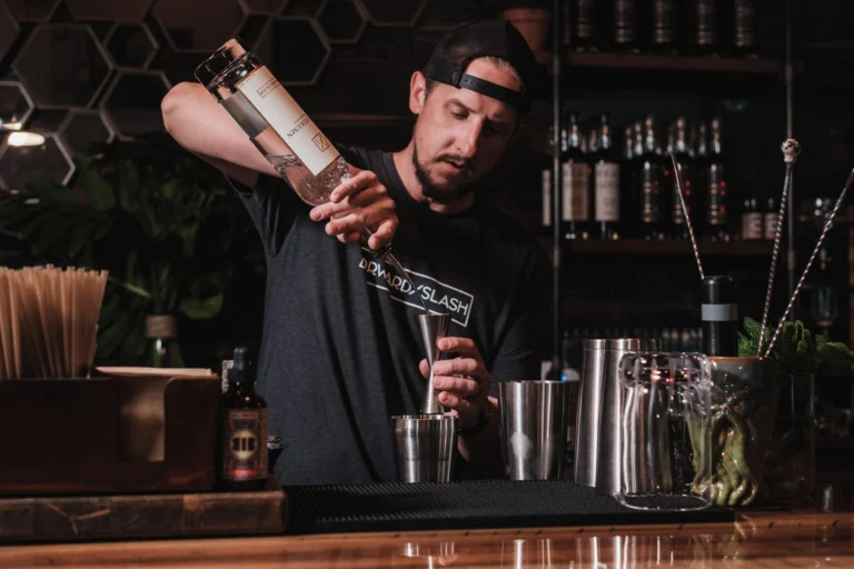 A male bartender carefully pouring alcohol into a shaker, preparing a cocktail at a bar counter.