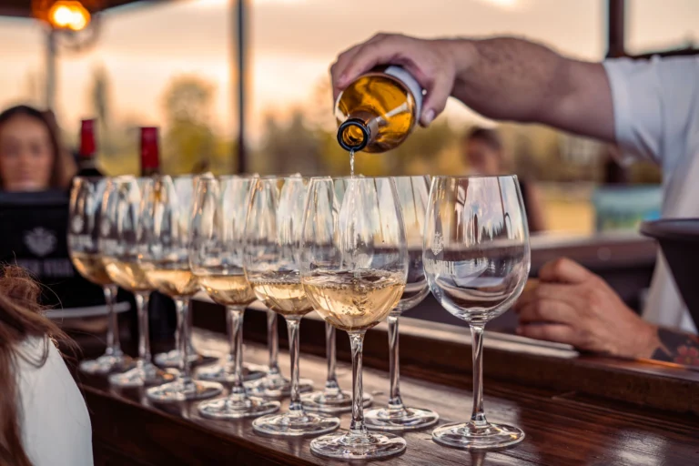 A barman pours white wine into glasses on a wooden counter at sunset.