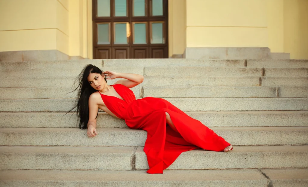 Stylish woman in a red gown lounging gracefully on steps indoors.