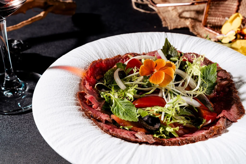 A fresh and vibrant gourmet beef carpaccio dish with leafy greens and edible flower garnish.