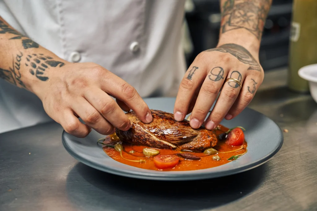 Chef with tattoos presenting a perfectly cooked steak with garnishes on a colorful plate.