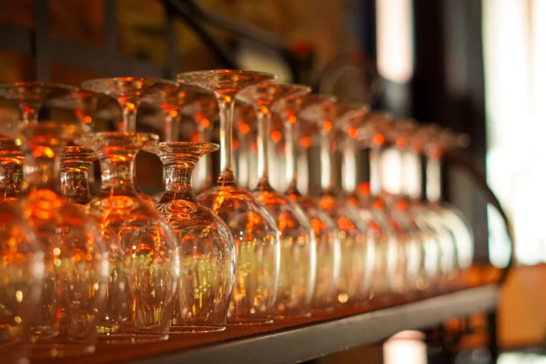 A close-up photo of upside-down wine glasses in a dimly lit bar, emphasizing light reflections and patterns.
