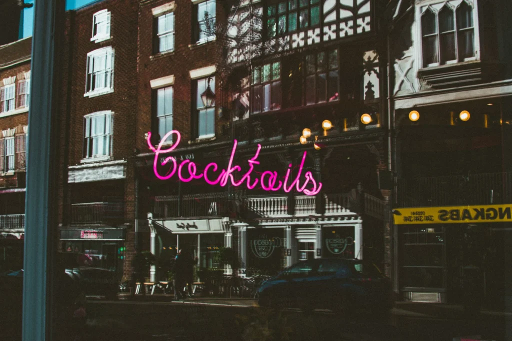 Pink neon cocktail sign reflected in urban architectural window at night.
