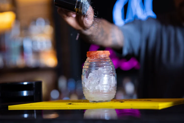Close-up of a bartender sprinkling spice on a cocktail in a vibrant bar environment.