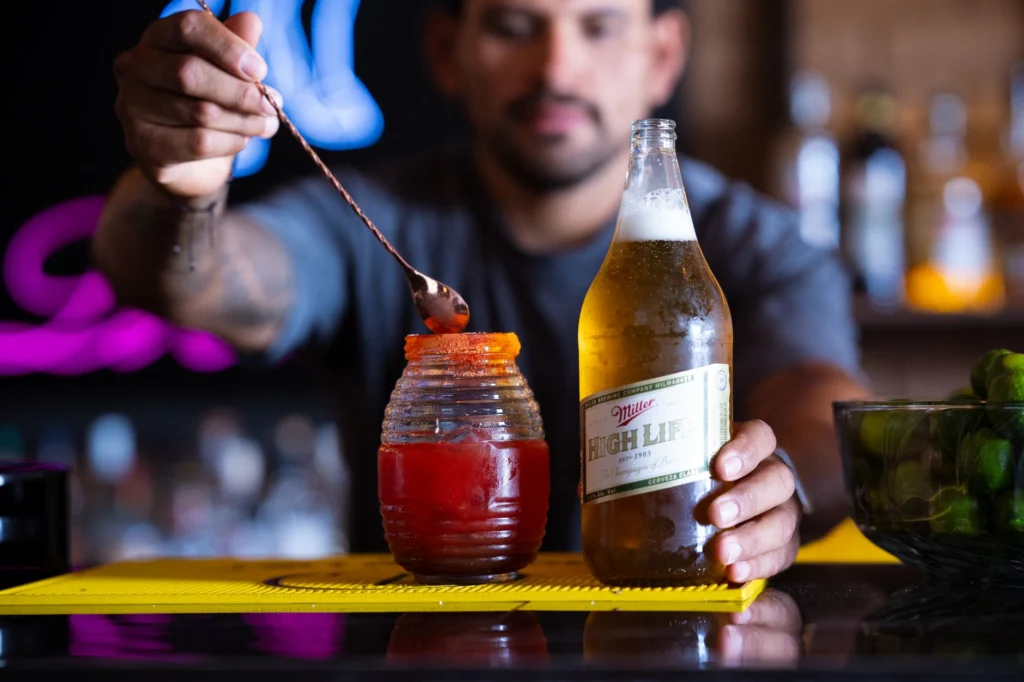 A bartender expertly prepares a michelada using Miller High Life beer in a vibrant bar setting.