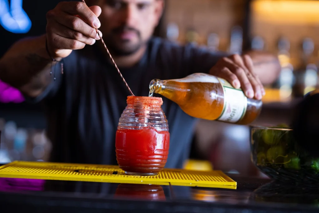 A bartender skillfully prepares a refreshing michelada with beer and tomato juice in a lively bar setting.