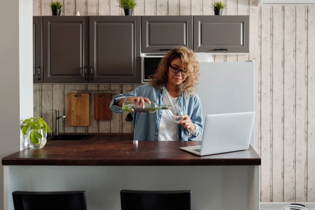 Woman pours wine while working on a laptop in her modern kitchen at home.