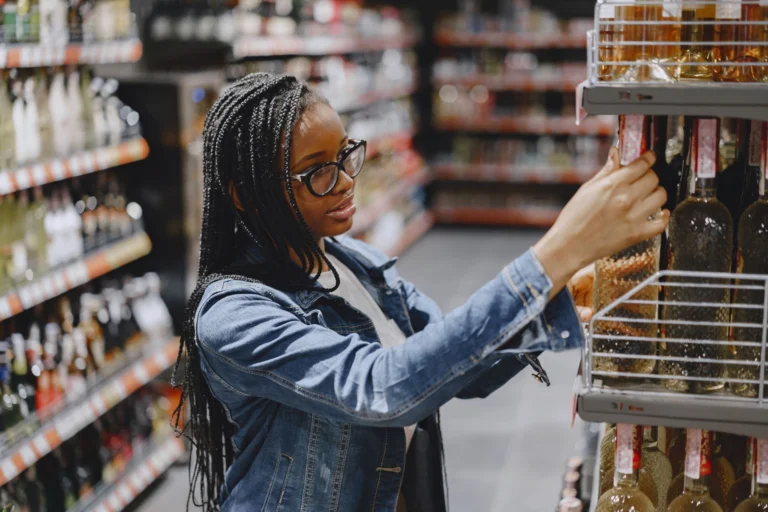A woman selects bottles of wine from a store shelf, shopping indoors.