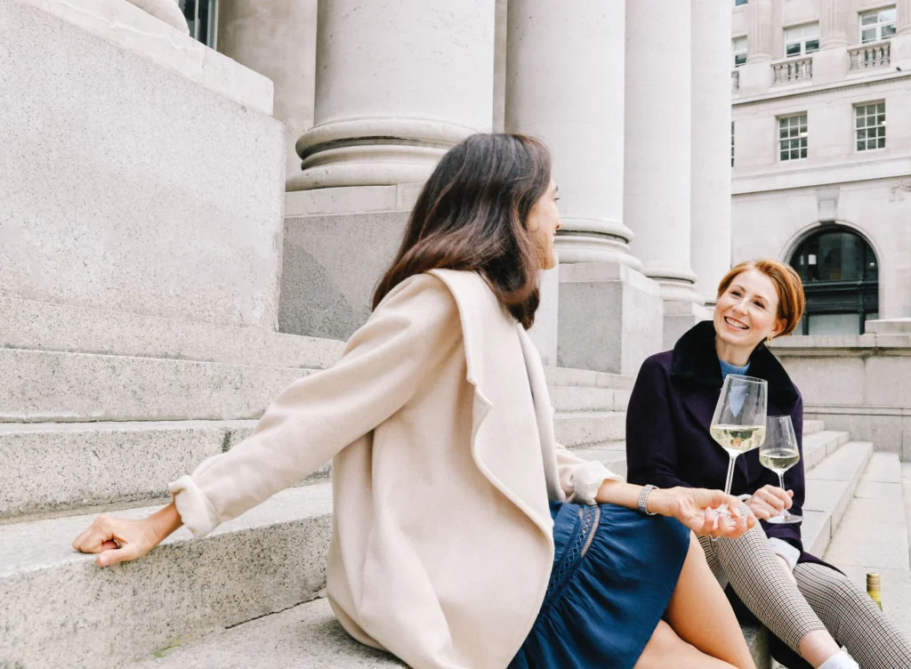 Two women enjoying a chat and wine on the steps of a historic London building on a clear day.