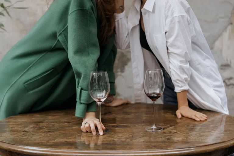 Two adults leaning over a wooden table with wine glasses, sharing a secret. Cozy and intimate atmosphere.