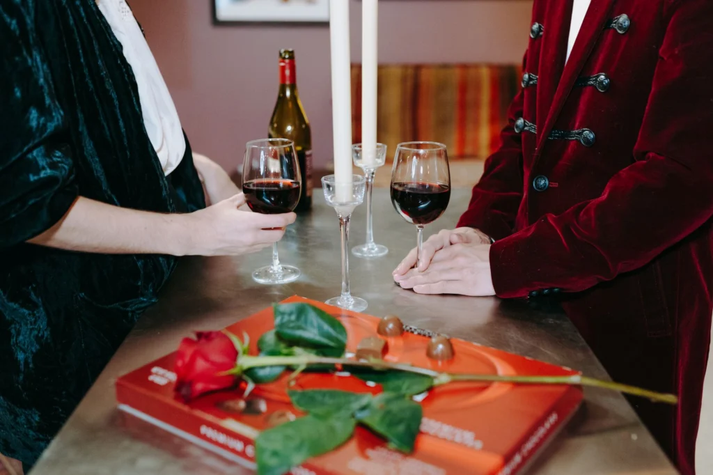 A couple enjoys red wine and chocolates by candlelight, with a red rose centerpiece.