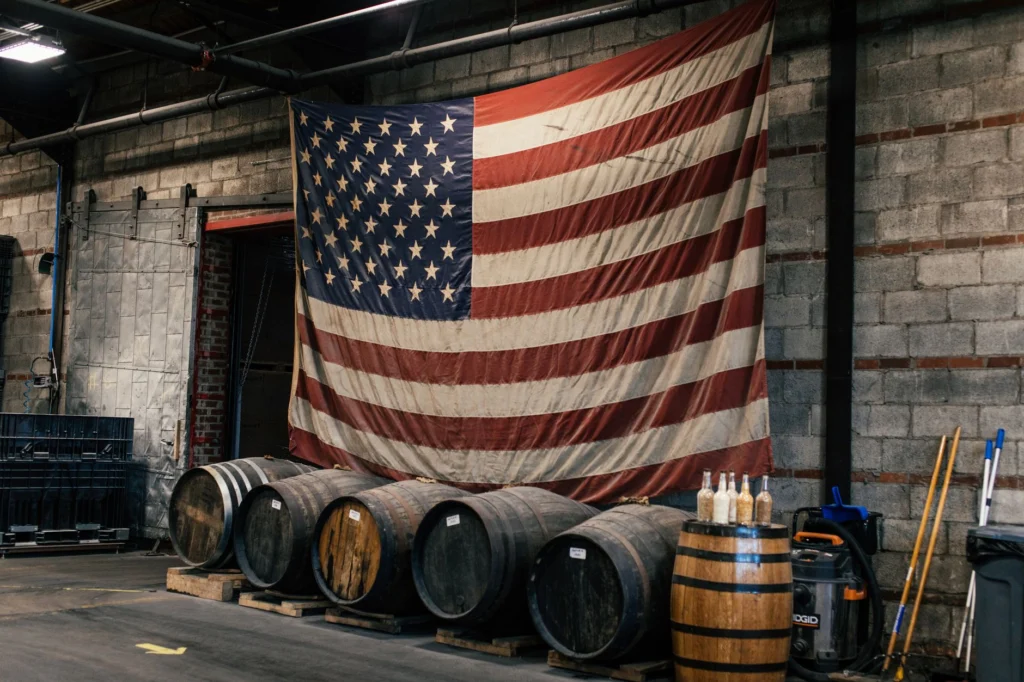 Indoor scene with American flag and wooden barrels in a rustic distillery setting.