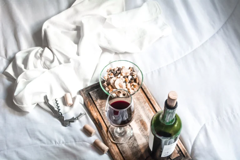 Aerial view of red wine, corkscrew, and nuts on wooden board on white bedding.