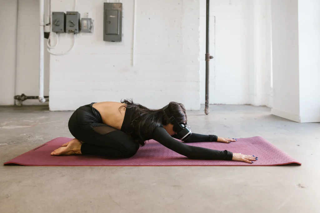 Adult woman using VR headset while performing yoga on a mat in a minimalist indoor setting.