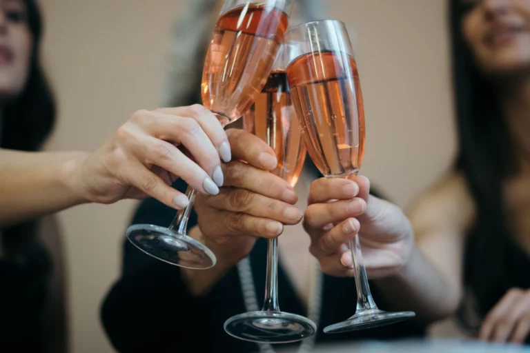 Close-up of four hands toasting with rosé wine glasses in a festive indoor setting.